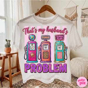 May include: White t-shirt with a graphic design featuring three colorful gas pumps. The text "That's my husband's PROBLEM" is printed below the pumps. The pumps are pink, orange, and teal.