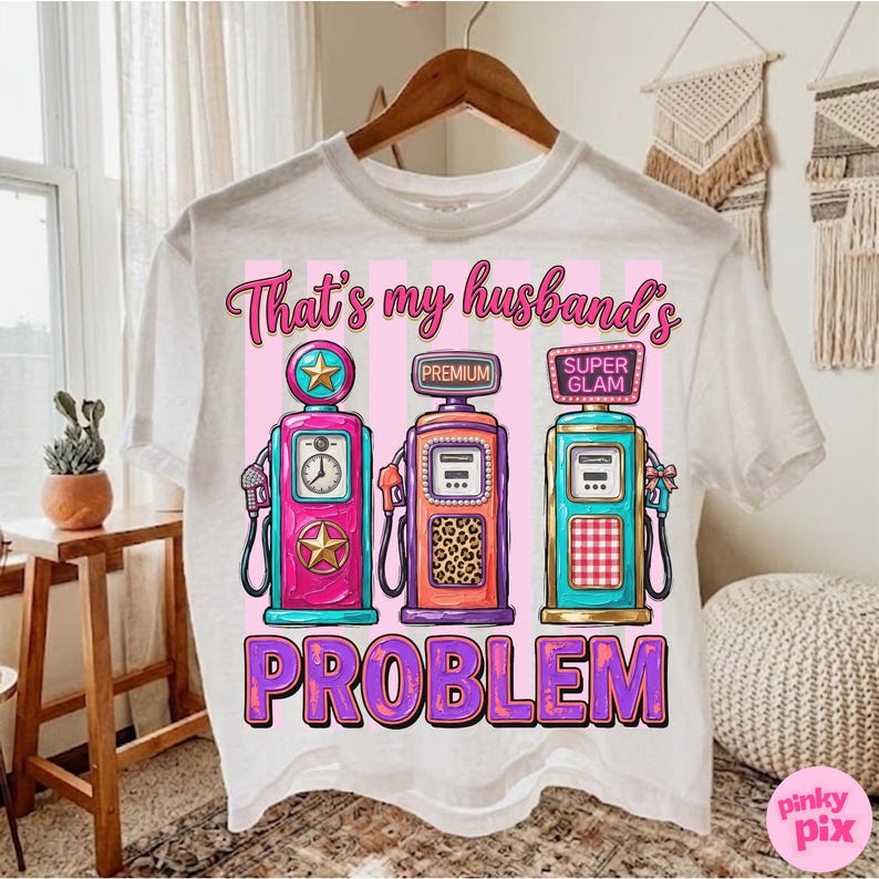 May include: White t-shirt with a graphic design featuring three colorful gas pumps. The text "That's my husband's PROBLEM" is printed below the pumps. The pumps are pink, orange, and teal.