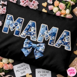 Puede incluir: Cojín negro con la palabra "MAMA" en letras de patchwork floral azul, contorneadas con perlas blancas. Un lazo floral azul a juego está debajo de la palabra. Las palabras "Happy Mother's Day" y "My Dear Mom" están en pequeñas tarjetas blancas.