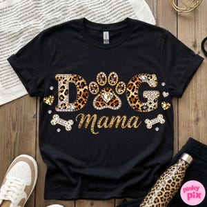 May include: Black t-shirt with the text "DOG Mama" in a leopard print design. The letters are embellished with rhinestones. The paw print in the center has a heart-shaped gem. The shirt also features bone and heart accents.