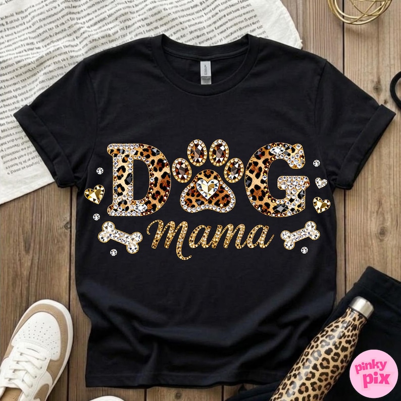 May include: Black t-shirt with the text "DOG Mama" in a leopard print design. The letters are embellished with rhinestones. The paw print in the center has a heart-shaped gem. The shirt also features bone and heart accents.