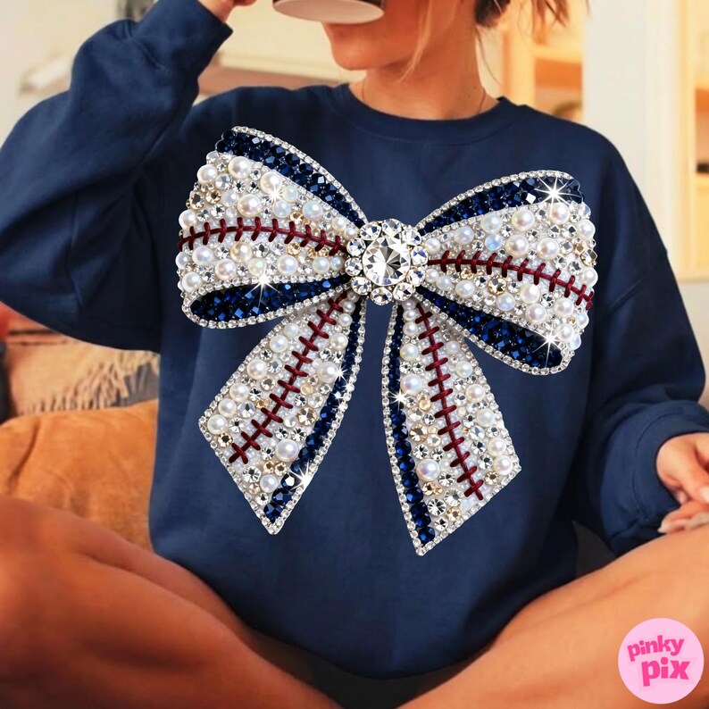 May include: Navy blue sweatshirt featuring a large bow design. The bow is embellished with white pearls, clear rhinestones, and dark blue and red accents. A large, clear rhinestone is in the center of the bow.