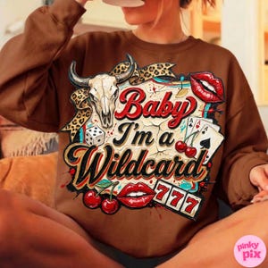 Może przedstawiać: Brązowa bluza z nadrukiem przedstawiającym czaszkę byka, karty do gry, wiśnie, kostki do gry oraz napis "Baby I'm a Wildcard". Projekt zawiera czerwone usta i liczbę 777.
