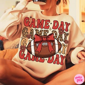 May include: Cream-colored sweatshirt with the words "GAME DAY" in red, leopard print, and floral accents. A brown football with a red bow is the central graphic. The sweatshirt is a casual, comfortable style.