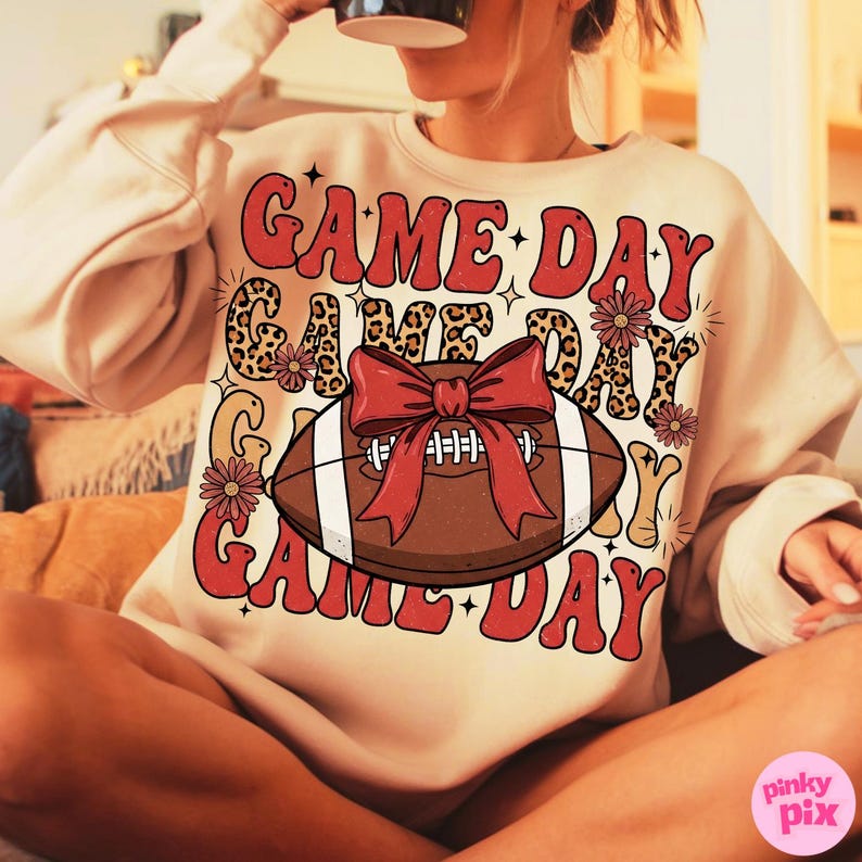 May include: Cream-colored sweatshirt with the words "GAME DAY" in red, leopard print, and floral accents. A brown football with a red bow is the central graphic. The sweatshirt is a casual, comfortable style.