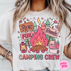 May include: Beige t-shirt with a colorful camping-themed graphic. The design includes a campfire, trees, and signs that read "Fires," "Fun," and "Friends." The words "Camping Crew" are also displayed.
