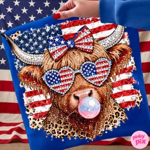 May include: Blue t-shirt featuring a Highland cow with patriotic embellishments. The cow wears heart-shaped sunglasses and a bow, both adorned with the American flag design. A bubble gum bubble adds a playful touch. The design incorporates leopard print and rhinestones.