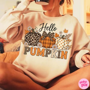 Op de afbeelding: Een beige sweatshirt met een herfstthema. De afbeelding toont drie pompoenen met verschillende patronen: luipaardprint, ruitjes en stippen. De tekst "Hello PUMPKIN" is gedrukt met bloemen- en bladeraccenten.