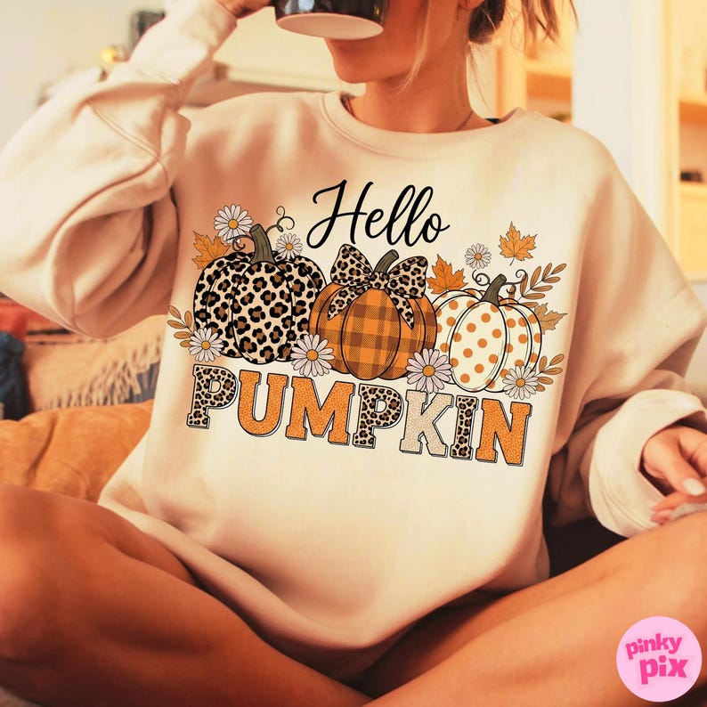 Op de afbeelding: Een beige sweatshirt met een herfstthema. De afbeelding toont drie pompoenen met verschillende patronen: luipaardprint, ruitjes en stippen. De tekst "Hello PUMPKIN" is gedrukt met bloemen- en bladeraccenten.