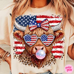 May include: A beige t-shirt featuring a graphic of a Highland cow with patriotic sunglasses and a bow, both adorned with the American flag. The cow is blowing a bubblegum bubble. The design includes a leopard print pattern.