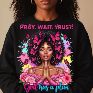 May include: Black sweatshirt featuring a vibrant graphic of a woman with pink butterflies and the text "PRAY. WAIT. TRUST." above her. Below the woman is the text "God has a plan". The design includes flowers and colorful splashes.