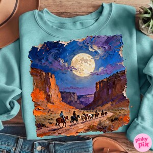 May include: A teal sweatshirt featuring a vibrant, impressionistic painting of cowboys riding through a canyon under a full moon. The artwork showcases a night sky with a large, bright moon and colorful clouds. The design is a unique blend of art and apparel.
