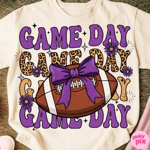 May include: Cream-colored t-shirt with a football graphic and the words "GAME DAY" in purple, leopard print, and orange. A purple bow is on the football, and small purple flowers are scattered around the design.