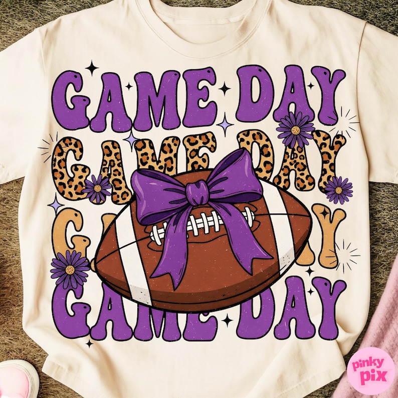 May include: Cream-colored t-shirt with a football graphic and the words "GAME DAY" in purple, leopard print, and orange. A purple bow is on the football, and small purple flowers are scattered around the design.