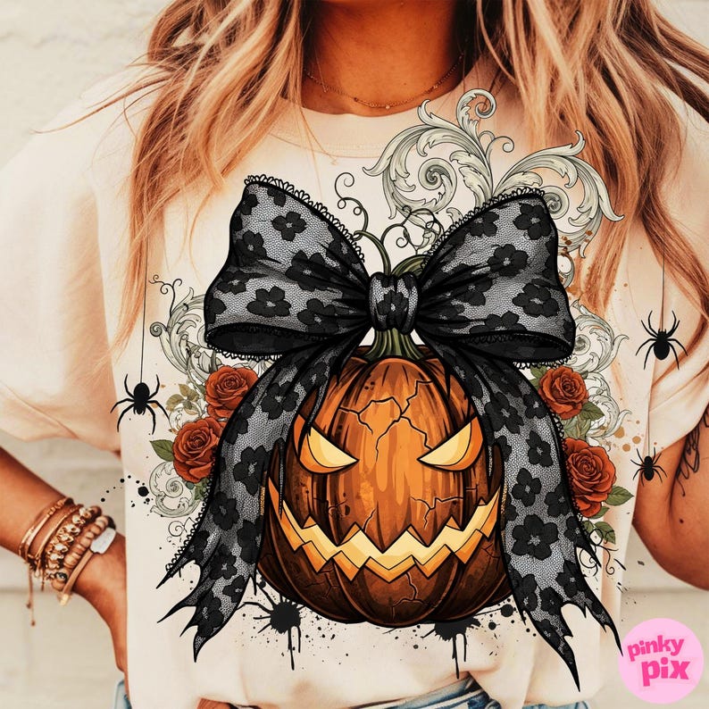 May include: A beige t-shirt featuring a Halloween-themed graphic. The design includes a carved orange pumpkin with a black lace bow, red roses, and spider accents. The text "pinky pix" is visible in the lower right corner.
