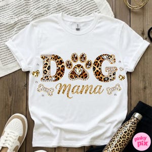 May include: White t-shirt with the text "DOG Mama" in a leopard print design, embellished with rhinestones and heart accents. The paw print in the center is also leopard print. A bone and heart accents are also included.