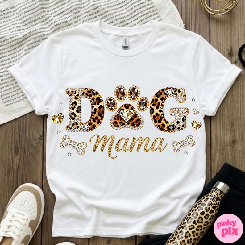 May include: White t-shirt with the text "DOG Mama" in a leopard print design, embellished with rhinestones and heart accents. The paw print in the center is also leopard print. A bone and heart accents are also included.