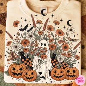 May include: A cream-coloured sweatshirt with a Halloween design. The graphic shows a ghost holding a pumpkin and flowers, with pumpkins, black cats, bats, and floral elements in orange, white, and black.