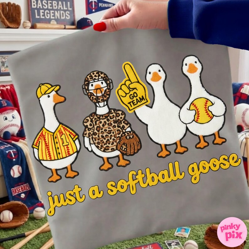 May include: A gray pillowcase featuring four cartoon geese dressed in baseball attire. One goose wears a baseball jersey, another a leopard print outfit, and a third holds a "Go Team" foam finger. The phrase "just a softball goose" is written in yellow.