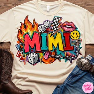 May include: Cream-colored t-shirt featuring a vibrant graphic design with the word "MIMI" in colorful graffiti-style lettering. The design includes flames, dice, a lightning bolt, lips, a smiley face, and barbed wire. The shirt is paired with blue jeans and a brown cowboy boot.