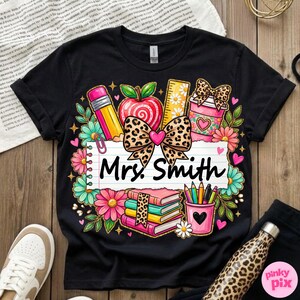 May include: Black t-shirt with a colorful teacher-themed graphic. The design includes a pencil, apple, ruler, books, and a coffee cup. The name "Mrs. Smith" is written on a lined paper. Floral and heart accents complete the design.