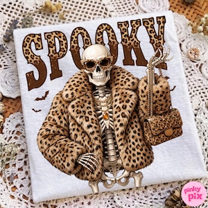 May include: A white t-shirt featuring a graphic of a skeleton wearing a leopard print faux fur coat, cat-eye sunglasses, and a gold chain necklace. The skeleton holds a quilted leopard print handbag. The word SPOOKY is printed in leopard print.