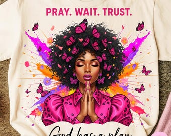 May include: Cream-colored t-shirt featuring a vibrant illustration of a woman with closed eyes, adorned with pink butterfly accents. The text "PRAY. WAIT. TRUST." and "God has a plan" are displayed in pink and black, respectively.