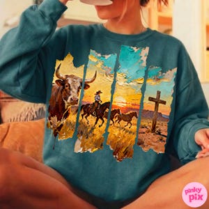 May include: Teal-colored sweatshirt featuring a Western-themed graphic. The design includes a bull, a cowboy on horseback, and a cross against a sunset backdrop. The artwork has a textured, painted appearance.
