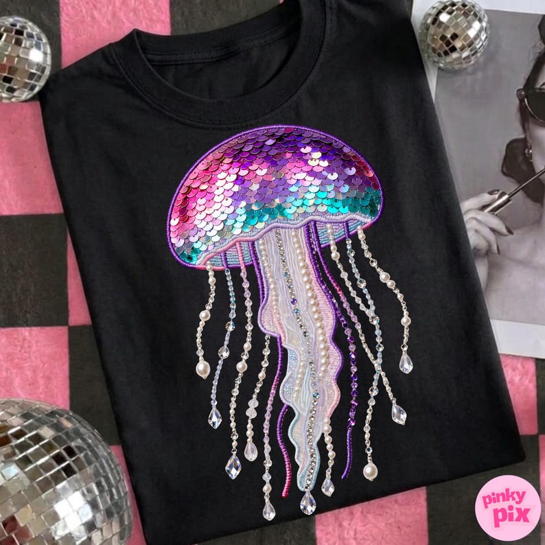 May include: Black t-shirt featuring a sequined jellyfish design in shades of pink, purple, and teal. The jellyfish has beaded tentacles with pearl and crystal accents. The shirt is a crew neck.