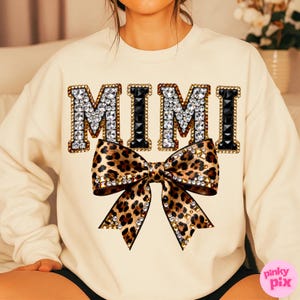 May include: Cream-colored sweatshirt with the word "MIMI" in large, jeweled letters. A leopard print bow with rhinestone accents is centered below the text. The sweatshirt is a casual style.