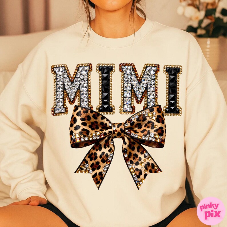 May include: Cream-colored sweatshirt with the word "MIMI" in large, jeweled letters. A leopard print bow with rhinestone accents is centered below the text. The sweatshirt is a casual style.
