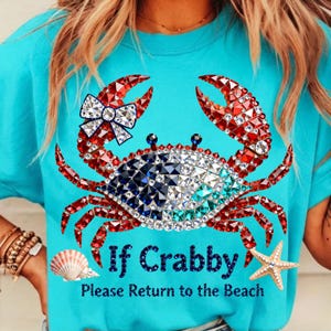May include: A turquoise t-shirt features a crab design made of red, white, blue, and clear rhinestones. The crab has a bow and the text "If Crabby Please Return to the Beach". A seashell and starfish are also on the shirt.