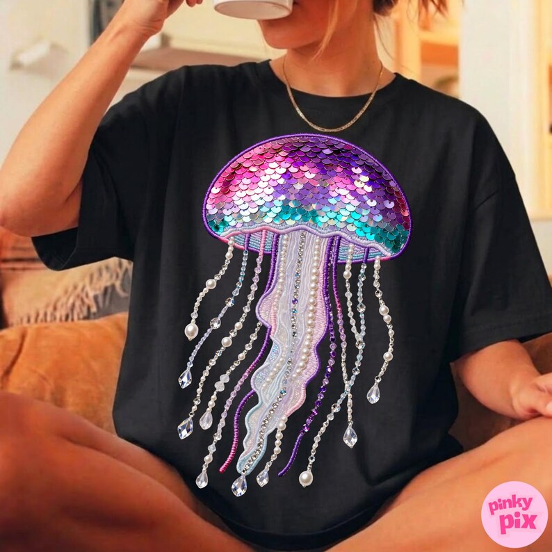 May include: Black t-shirt featuring a colorful jellyfish design. The jellyfish has a sequined dome in shades of pink, purple, and teal, with beaded and crystal-like strands. The design is centered on the shirt.