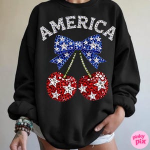 May include: Black sweatshirt with the word "AMERICA" in sparkling white letters. A blue bow with white stars sits above two red cherries with white star accents. The design is patriotic and festive.