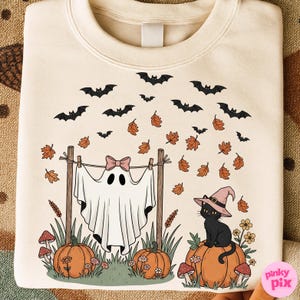 May include: Cream-colored sweatshirt with a Halloween-themed graphic. The design features a ghost hanging on a clothesline with a pink bow, pumpkins, a black cat wearing a witch hat, bats, and autumn leaves.