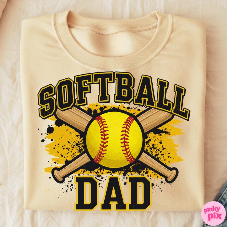 May include: Cream-colored sweatshirt with the words "SOFTBALL DAD" in black and yellow lettering. A yellow softball with red stitching is centered between two crossed wooden bats. The design includes yellow and black splatter effects.