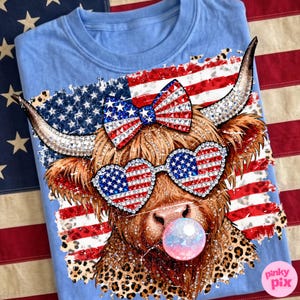 May include: Light blue t-shirt featuring a brown Highland cow with rhinestone accents, heart-shaped sunglasses, and a bow, all decorated with the American flag. The cow is blowing a bubble and has a leopard print collar. The shirt is on an American flag background.