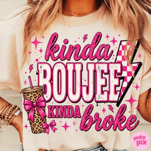 May include: A cream-colored t-shirt featuring a graphic print with the text "kinda BOUJEE kinda broke". The design includes a leopard print tumbler with a bright pink bow, a pink and white checkered lightning bolt, and various pink star icons.