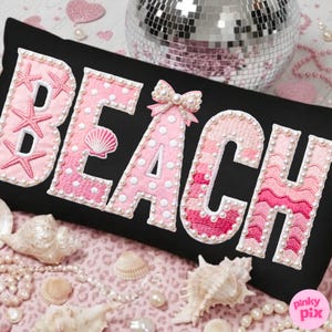 May include: Black rectangular pillow with the word "BEACH" embroidered in pink, adorned with pearls and seashell details. The letters are embellished with starfish, polka dots, and a bow. Decorative seashells and pearls surround the pillow.