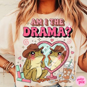 以下が含まれることがあります： ピンクと茶色で「AM I THE DRAMA？」と書かれたベージュのTシャツ。カウボーイハットをかぶった漫画のカエルが、ハート型の鏡を見ている様子が描かれています。口紅、デイジー、飲み物も表示されています。