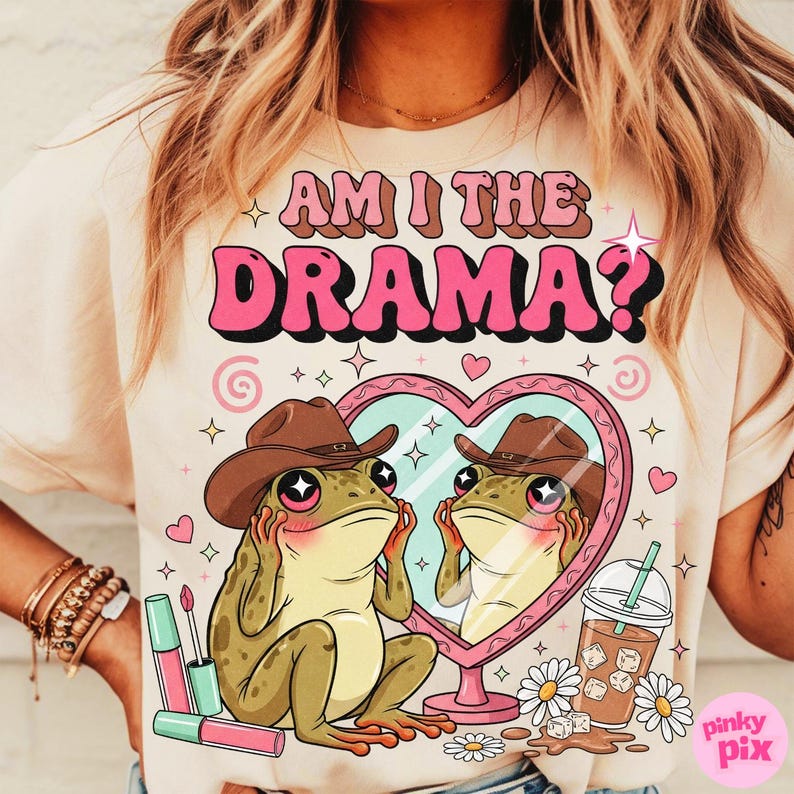 以下が含まれることがあります： ピンクと茶色で「AM I THE DRAMA？」と書かれたベージュのTシャツ。カウボーイハットをかぶった漫画のカエルが、ハート型の鏡を見ている様子が描かれています。口紅、デイジー、飲み物も表示されています。