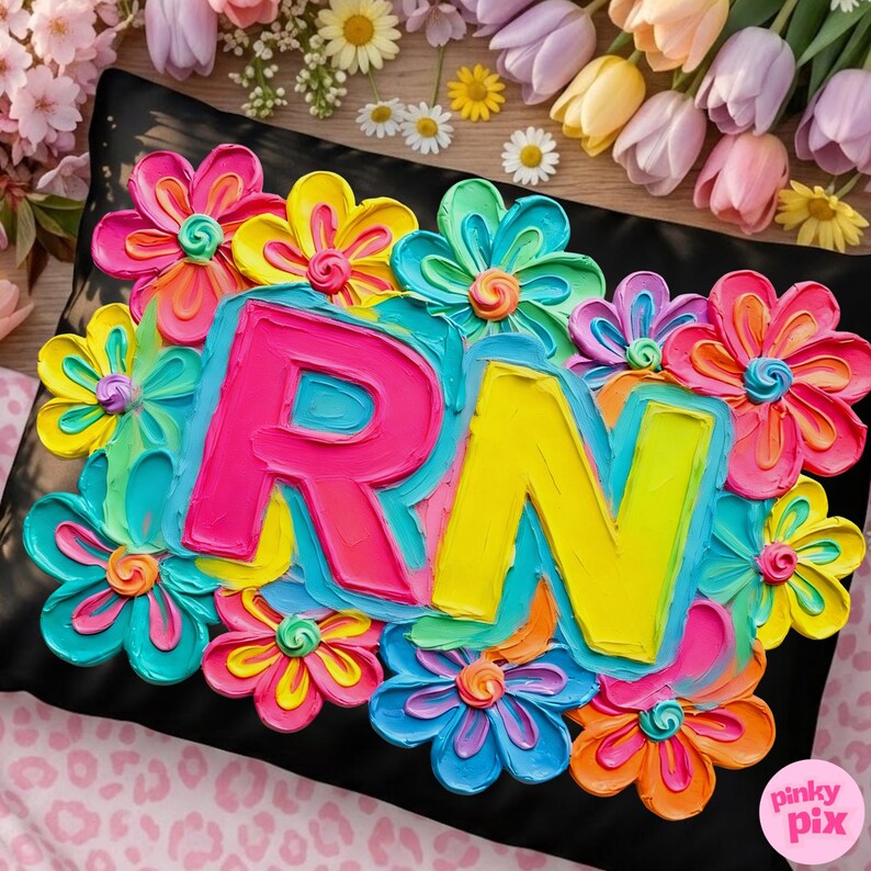 Op de afbeelding: Zwart kussen met kleurrijke, getextureerde bloemen en de letters "RN" in felroze en geel. De bloemen zijn in tinten roze, blauw, geel en oranje. Het kussen is omringd door bloemen.