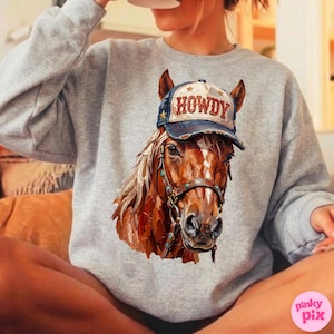 May include: A gray sweatshirt featuring a detailed illustration of a brown horse wearing a blue and white cap with the word "HOWDY". The horse has a bridle with turquoise accents. The sweatshirt is a casual style.