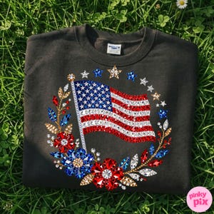 May include: Dark gray sweatshirt featuring a sparkling American flag design. The flag is embellished with red, white, and blue gemstones, surrounded by a wreath of flowers and stars. A patriotic apparel item.