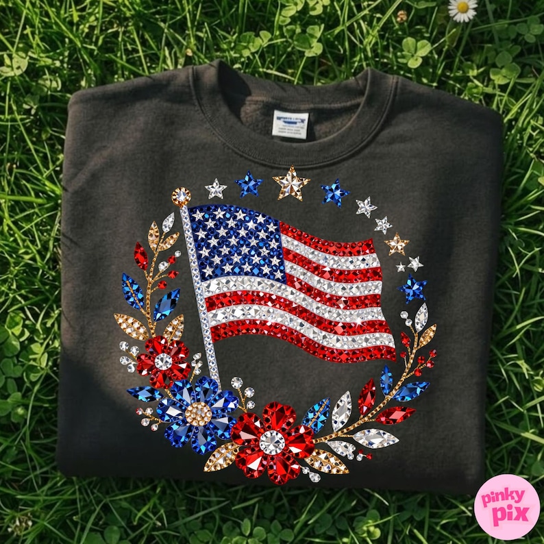 May include: Dark gray sweatshirt featuring a sparkling American flag design. The flag is embellished with red, white, and blue gemstones, surrounded by a wreath of flowers and stars. A patriotic apparel item.