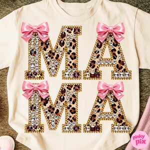May include: A cream-coloured t-shirt displaying the word "MAMA" in large, embellished letters. The letters have a leopard print design, gold-coloured trim, and sparkling rhinestones. Each letter is topped with a pink bow.