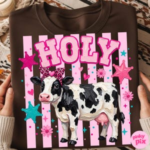 May include: Brown sweatshirt featuring a cow illustration with a leopard print bow and the word "HOLY" in pink letters. The design includes pink stripes, stars, and flowers. The cow is black and white.