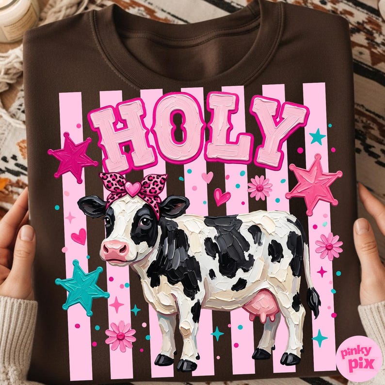 May include: Brown sweatshirt featuring a cow illustration with a leopard print bow and the word "HOLY" in pink letters. The design includes pink stripes, stars, and flowers. The cow is black and white.