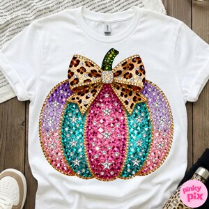 Op de afbeelding: Een wit t-shirt met een grote afbeelding van een pompoen versierd met roze, turquoise en paarse strass-steentjes en sterren. De pompoen is bekroond met een strik met luipaardprint, versierd met strass-steentjes en een groene steel.
