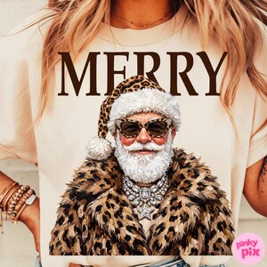 以下が含まれることがあります： クリーム色のTシャツで、茶色の文字でMERRYと書かれ、その下にヒョウ柄のサンタ帽子、ヒョウ柄のコート、大きなサングラス、大きなクリスタルの星のネックレスを身につけ、白いひげを生やした人物のグラフィックが描かれています。
